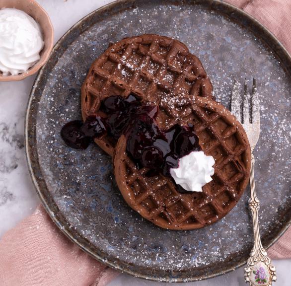 Zwei runde Schoko-Waffeln angerichtet auf einem Teller getoppt von Blaubeeren, Sauce und Sahne