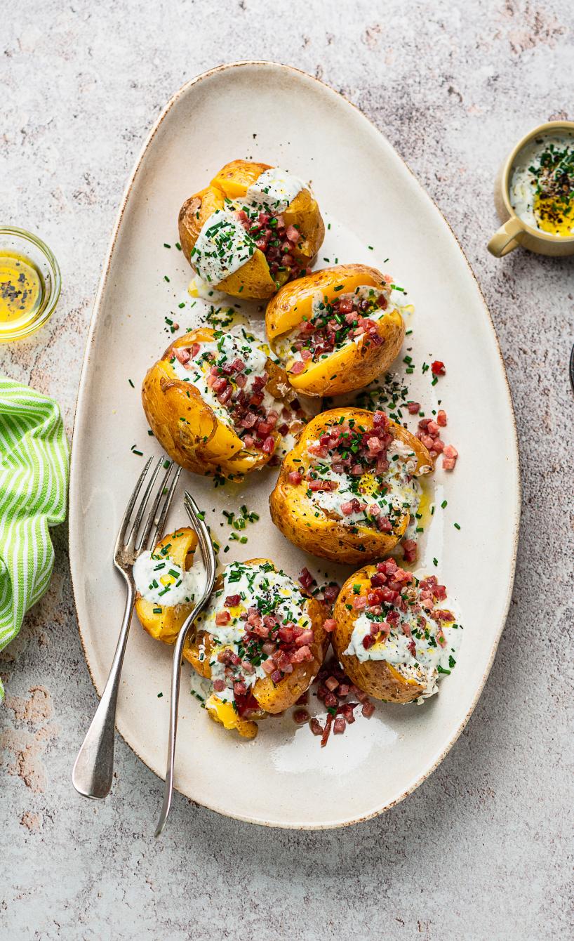 Eingeschnittene Ofenkartoffeln getoppt mit Schinkenwürfeln und Joghurtdip auf einem Teller