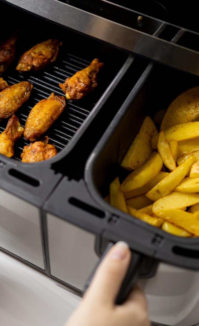 Eine Frau öffnet eine Heißluftfritteuse mit zwei Kammern, in der einen Chicken Wings, in der anderen Pommes