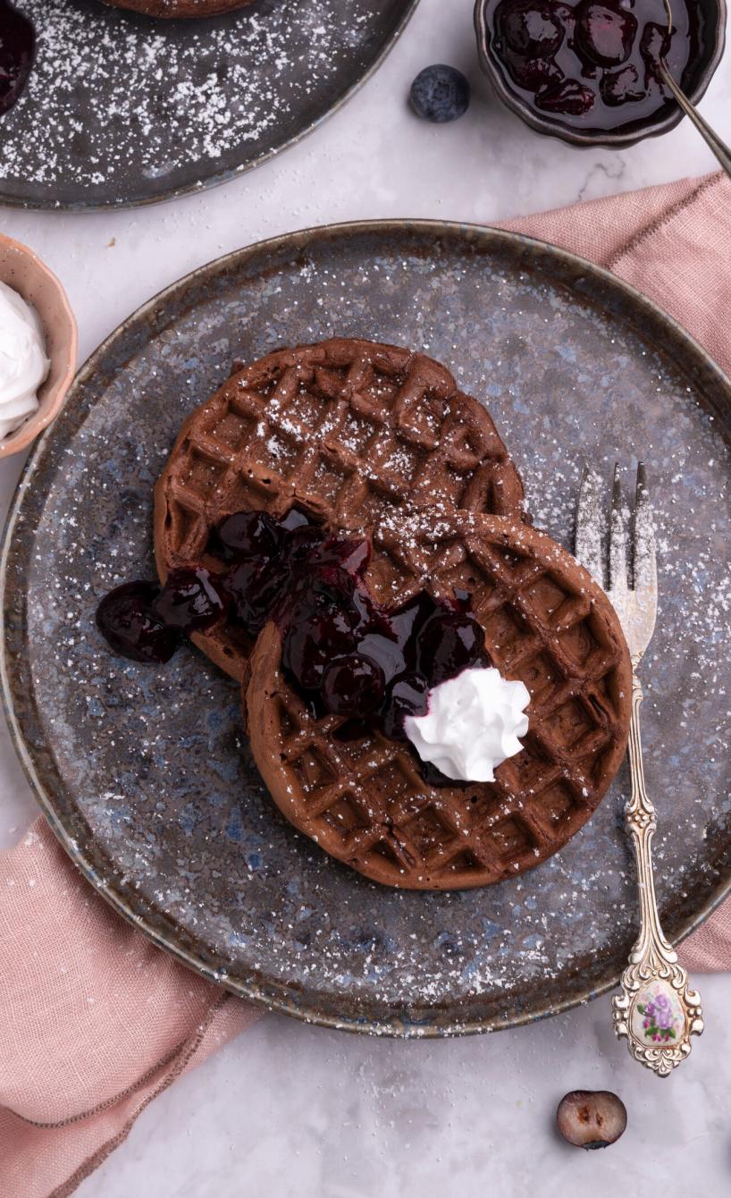 Zwei runde Schoko-Waffeln angerichtet auf einem Teller getoppt von Blaubeeren, Sauce und Sahne