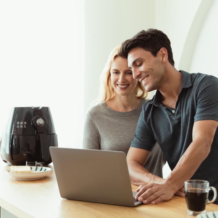 Pärchen steht an einer Kücheninsel und schaut sich Heißluftfritteusentipps auf dem Laptop an, daneben ein Airfryer
